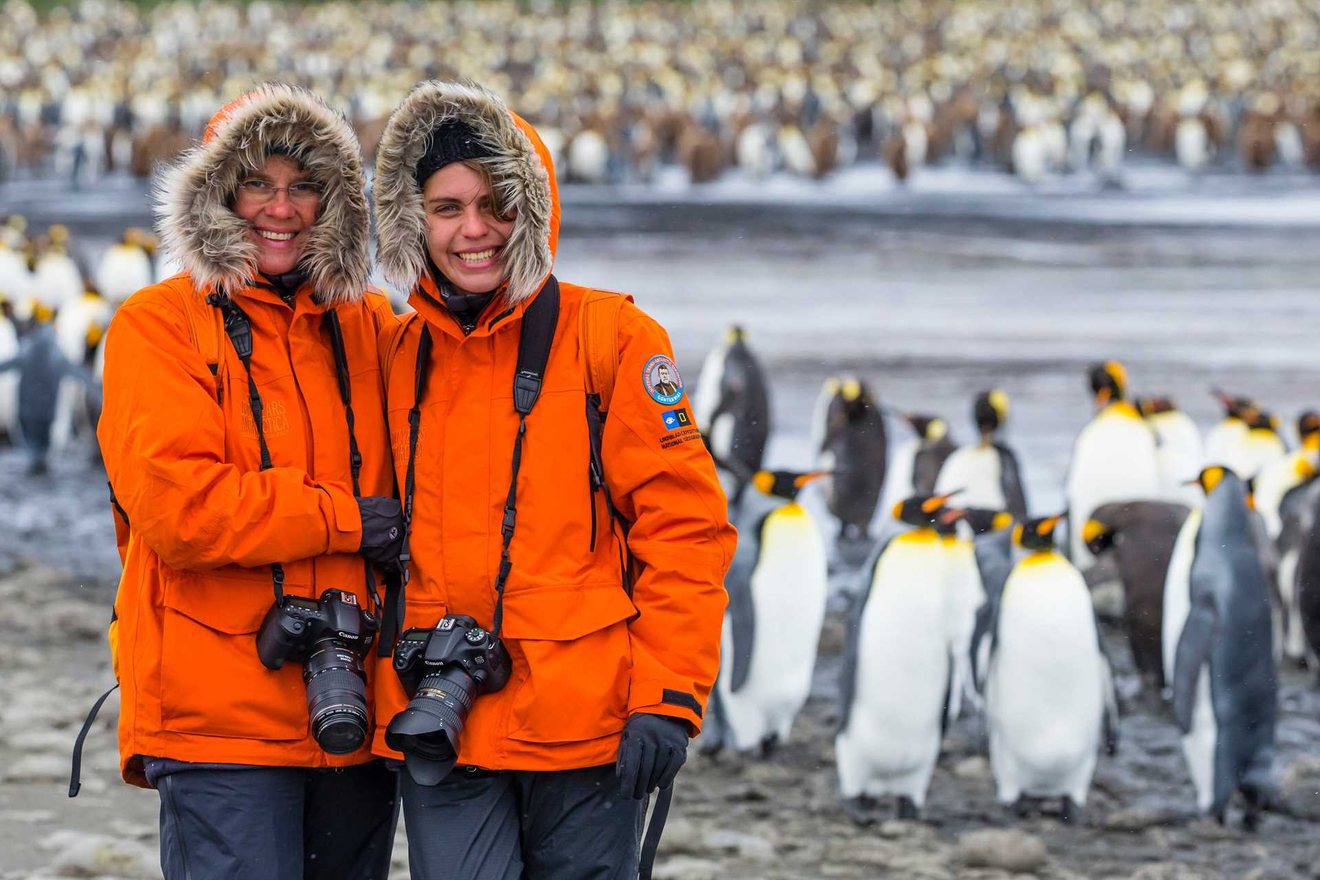 A mother and daughter in orange parkas pose in front of a large king penguin colony on Salisbury Plains, South Georgia.