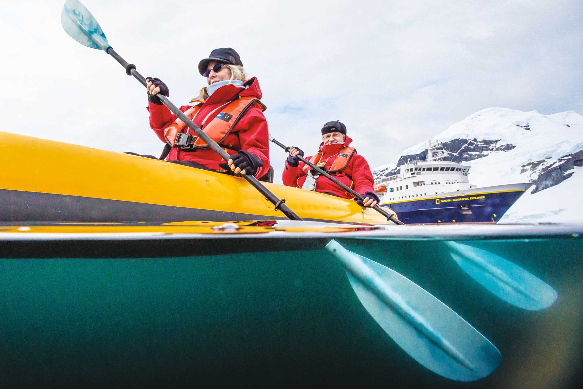 Two travelers kayak in the Errera Channel with the National Geographic Explorer in the distance.