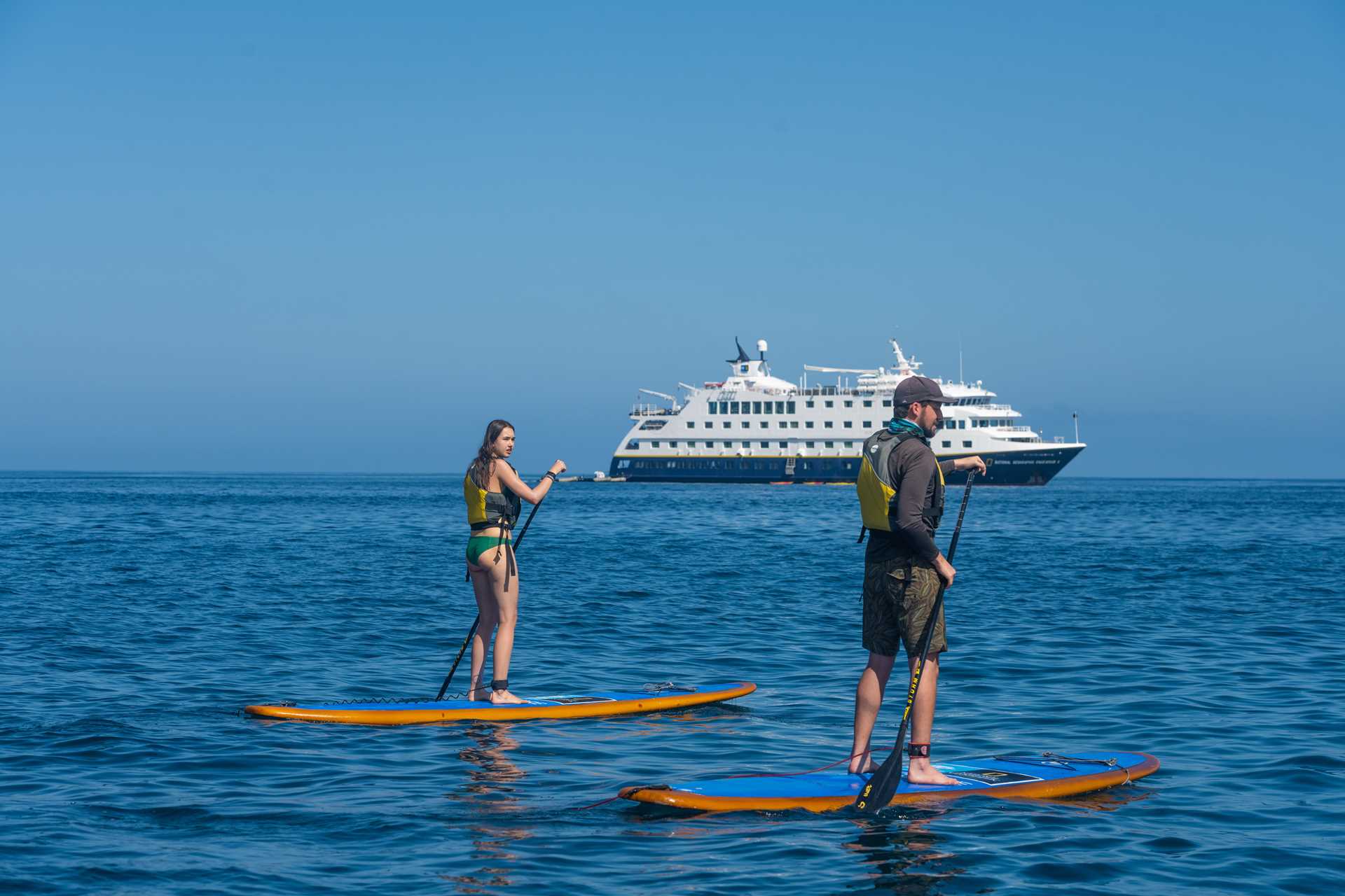 Travelers paddle on standup paddleboards off Santiago Island, Galápagos with the ship National Geographic Endeavour II in the distance.