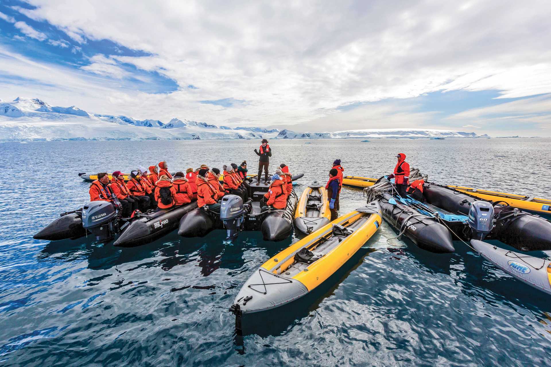 A naturalist standing on a Zodiac talks to a group of guests in multiple Zodiacs with kayaks tied off to them.