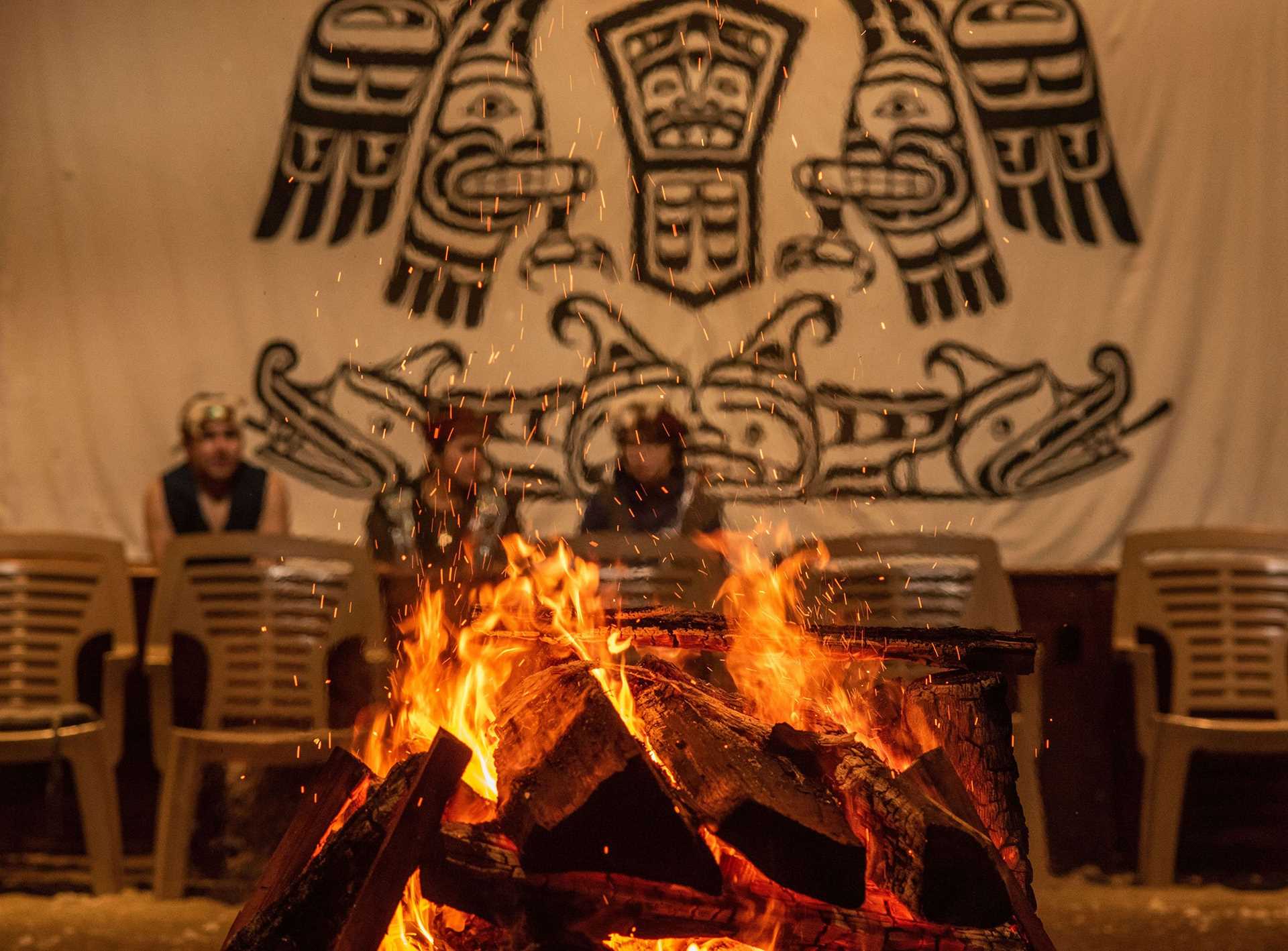 a fire inside the Alert Bay Big House, with a black and white wall mural visible in the background