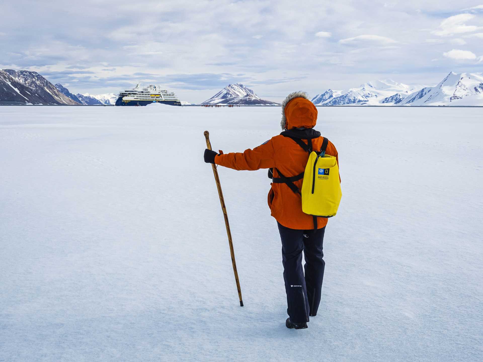 A guest hikes with a walking stick and yellow day pack towards the National Geographic Endurance in Bourgeois Fjord.