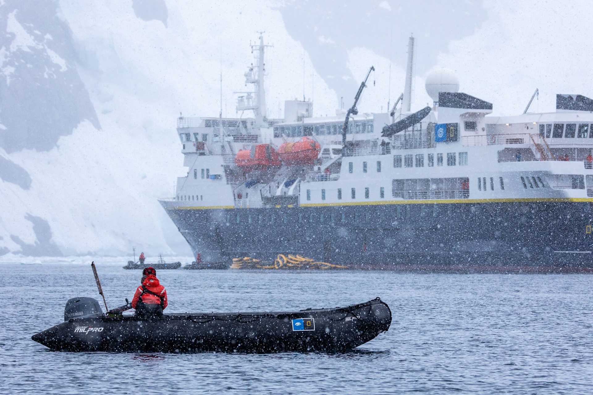 a zodiac craft in front of a larger ship, with snow falling all around them