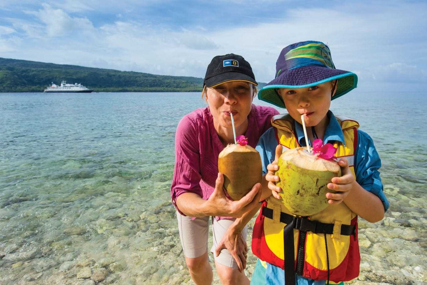 Two people&mdash;and adult and a child&mdash;drink out of coconuts using straws. In the background, a cruise ship sits in the bay