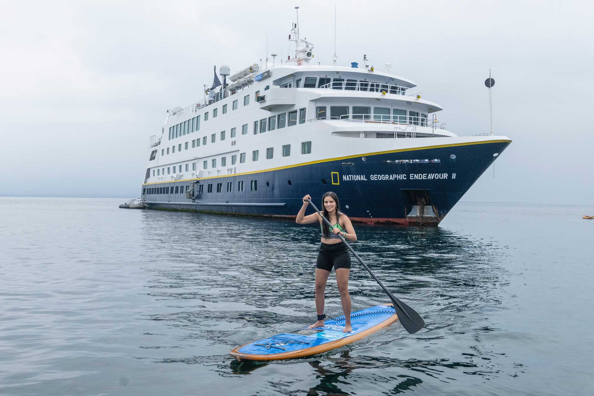 A guest stand-up paddleboards with the National Geographic Endeavour II in the background.